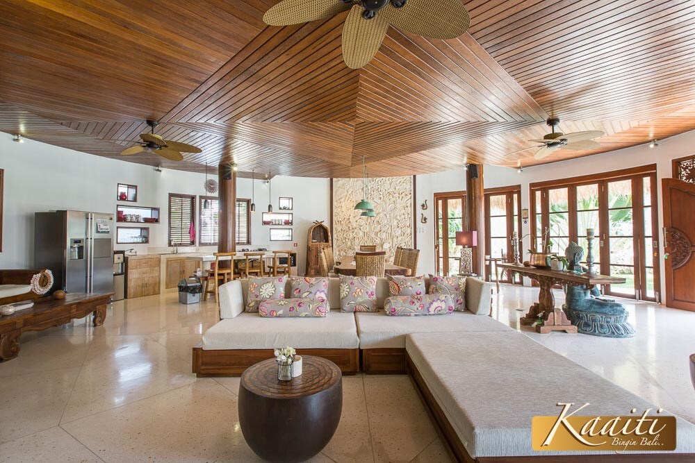 An open living area showcases a spacious layout with natural light filtering through large windows. A large, cushioned seating arrangement, adorned with decorative pillows, complements a round wooden table. Wooden beams and ceiling fans enhance the tropical aesthetic, while tasteful decor elements add character.