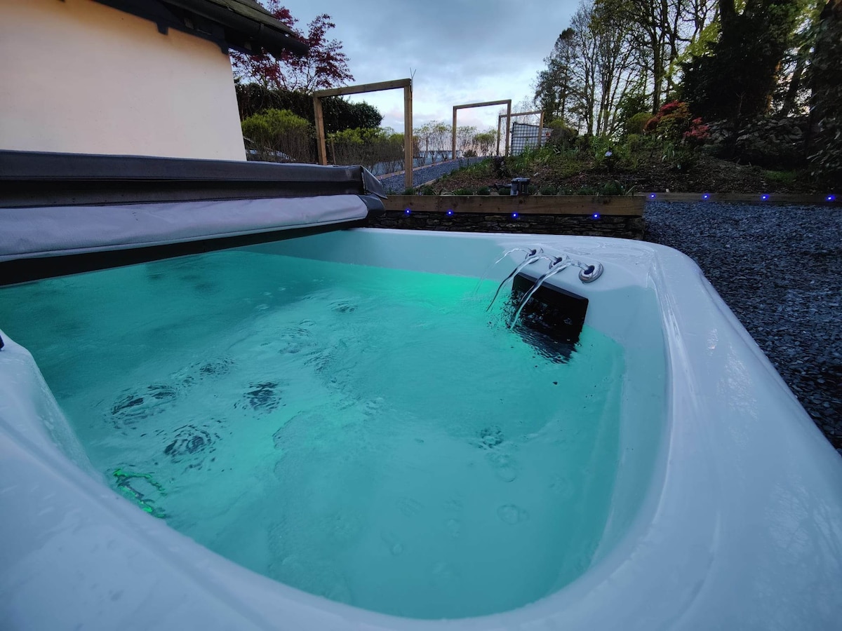 A private outdoor hot tub is set in a landscaped area, featuring illuminated water in a calming shade of green. Surrounding foliage and soft lighting create a tranquil ambiance for relaxation.