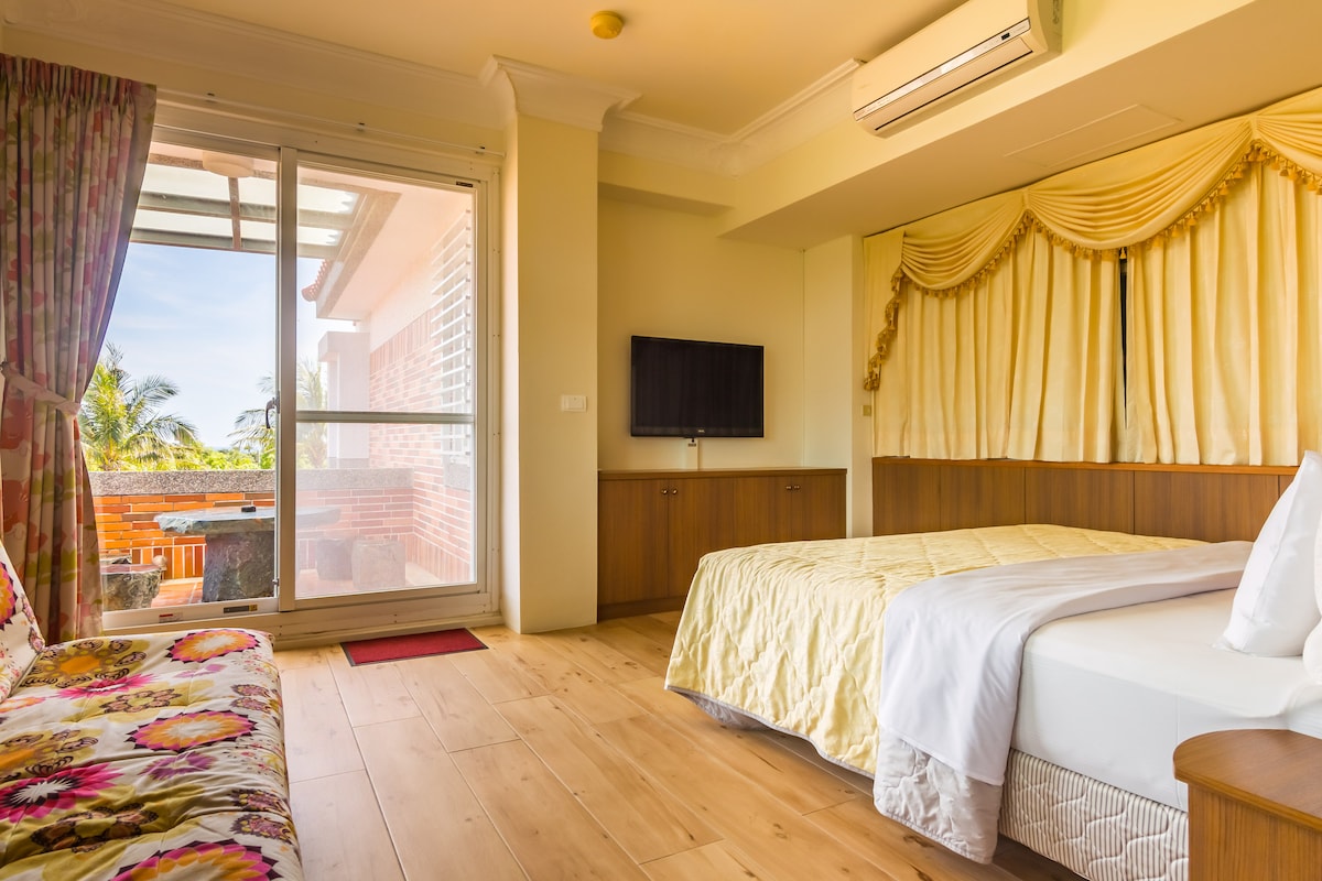 A bright room is showcased, featuring a spacious bed with light-colored bedding. A plush sofa adorned with floral patterns provides additional seating. Large glass doors lead to a balcony area, while a flat-screen TV is mounted on the wall opposite the bed.