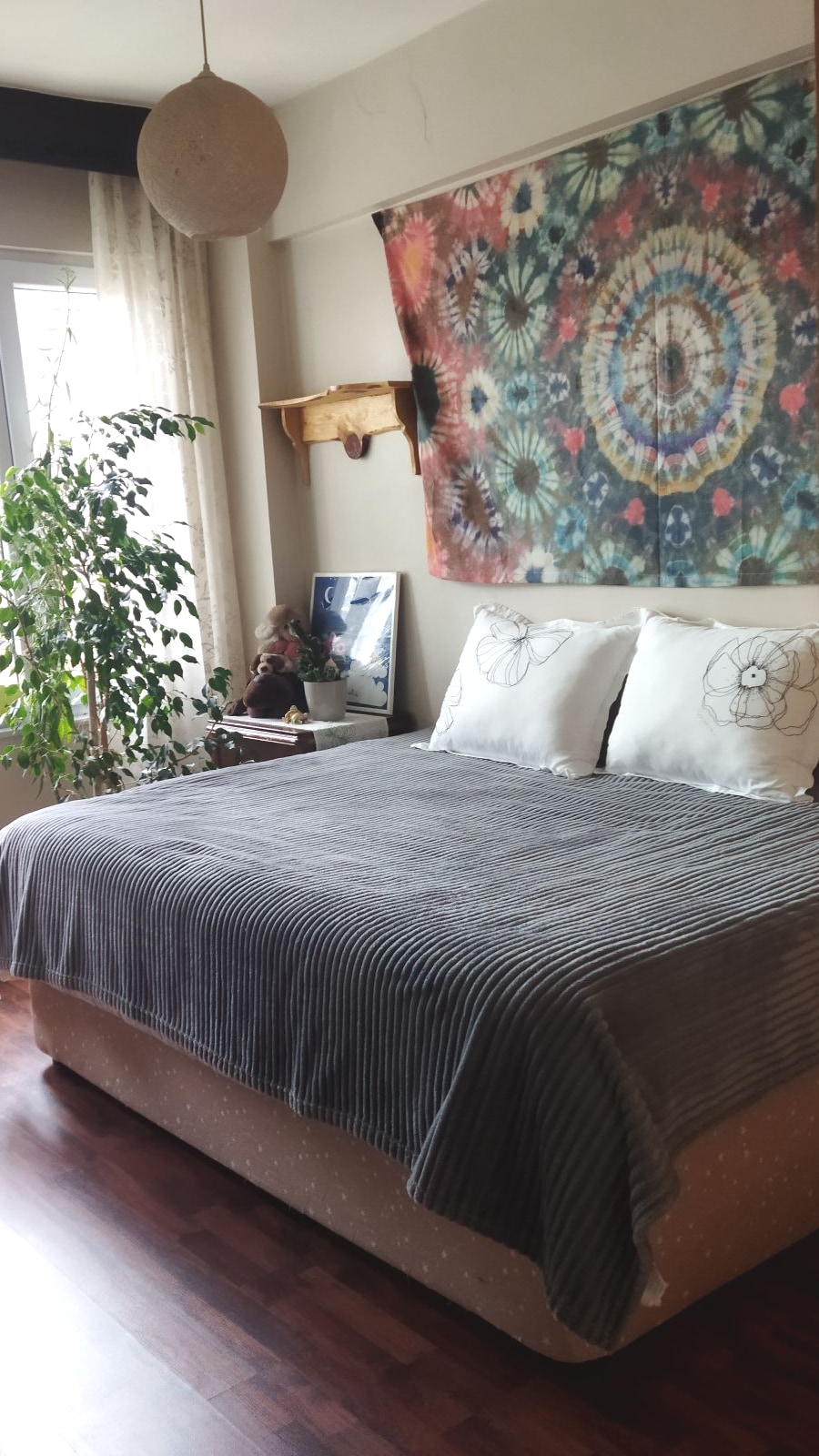 A spacious sleeping area features a large bed adorned with striped bedding and plush pillows. A colorful tapestry hangs above the bed, complementing the serene atmosphere. A tall plant is placed in the corner, with natural light filtering in through a nearby window, partially covered by light curtains.