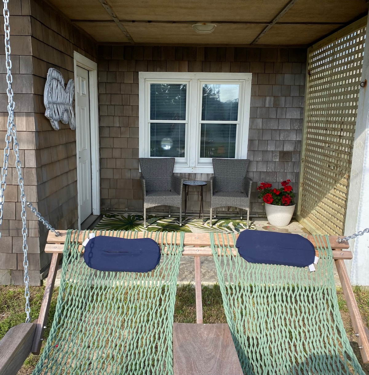 A welcoming outdoor area features two wicker chairs and a small table, set against a wooden shingle backdrop. A gently swaying hammock with cushions invites relaxation. A potted plant adds a touch of greenery, while sunlight filters through the nearby window.