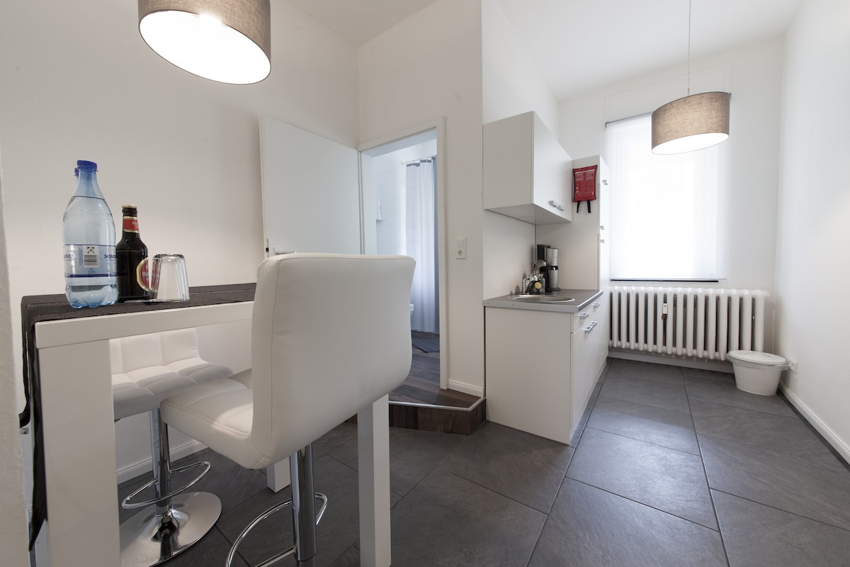 A modern kitchen area is presented, featuring sleek white cabinetry and a dark countertop. A small dining space with high chairs is visible. Natural light enters through a window. Appliances include a coffee maker and kettle, with a refrigerator located nearby.