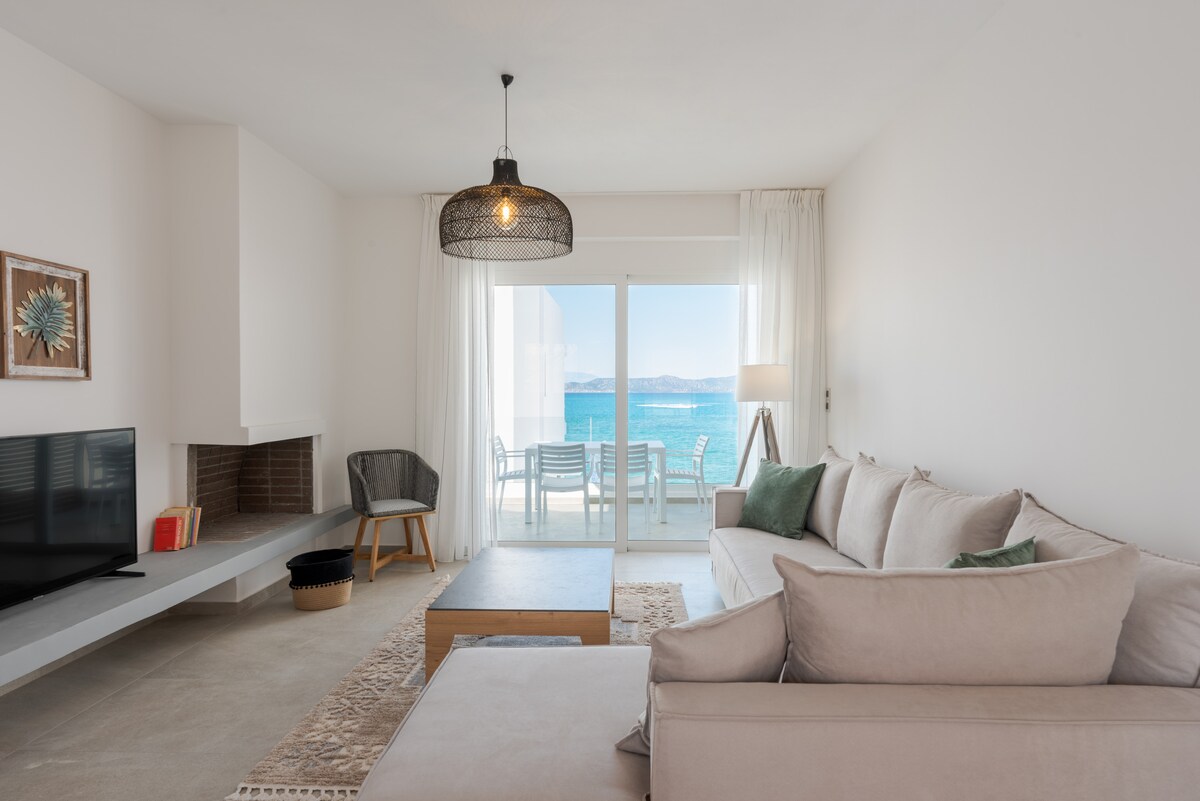 A light-filled living area features large glass doors that open to a balcony with sea views. A comfortable sofa and a minimalist coffee table complement the space. A television is positioned on a shelf near a simple fireplace, adding a touch of elegance.