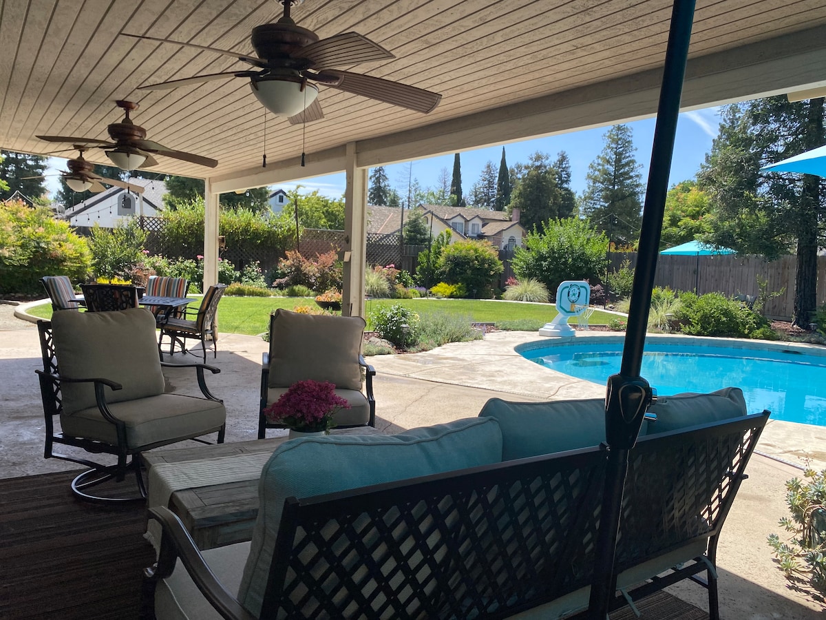 A covered patio area features comfortable seating arrangements, including soft chairs and a cozy couch. A dining table with colorful chairs is visible, alongside a glistening pool surrounded by landscaped gardens. Sunlight filters through the structure, creating a bright outdoor atmosphere.