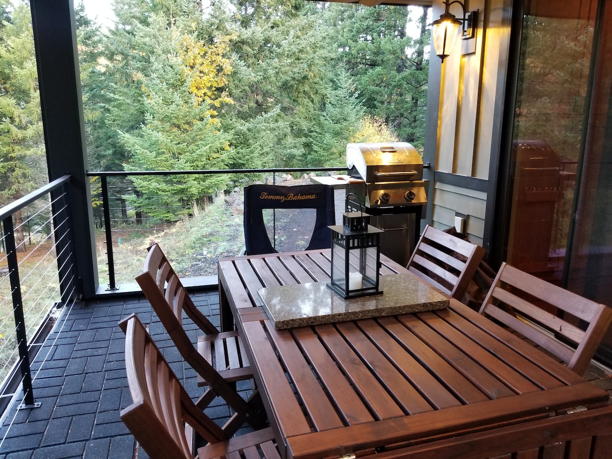A spacious outdoor deck is furnished with a large dining table made of wood, surrounded by several chairs. A grilling area is visible with a stainless steel barbecue. The background features a serene view of trees, providing a natural and secluded environment.