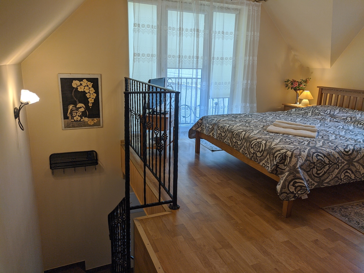 A well-lit bedroom is positioned on the upper level, featuring a spacious bed adorned with a patterned cover. A small balcony is visible through the large glass door, which is draped with sheer curtains. A bedside lamp and artwork complement the decor.