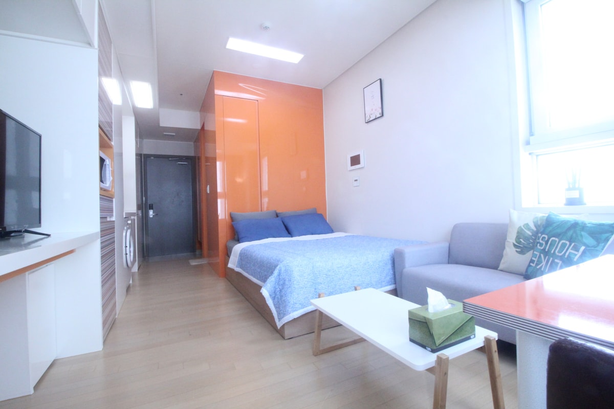 A bright studio space features a queen-sized bed with blue bedding. A gray sofa is positioned to the side, accompanied by a low coffee table. A TV is mounted on the wall, and large windows allow natural light to fill the room.