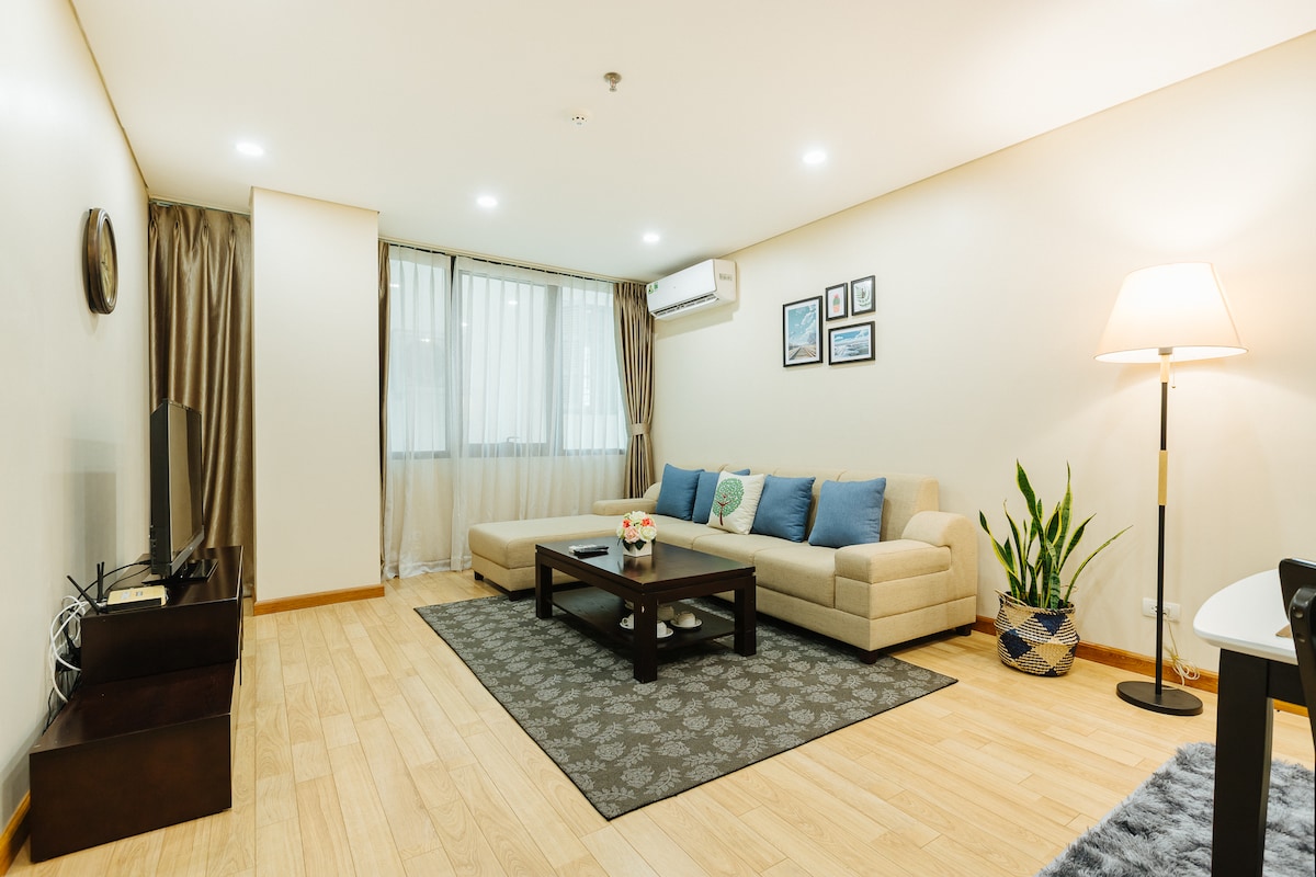 A spacious living area features light wood flooring and neutral-colored walls. A comfortable beige sofa is accompanied by blue accent pillows, while a dark coffee table sits at the center. Natural light filters through large windows, partially covered by sheer curtains, adding a bright touch to the room.