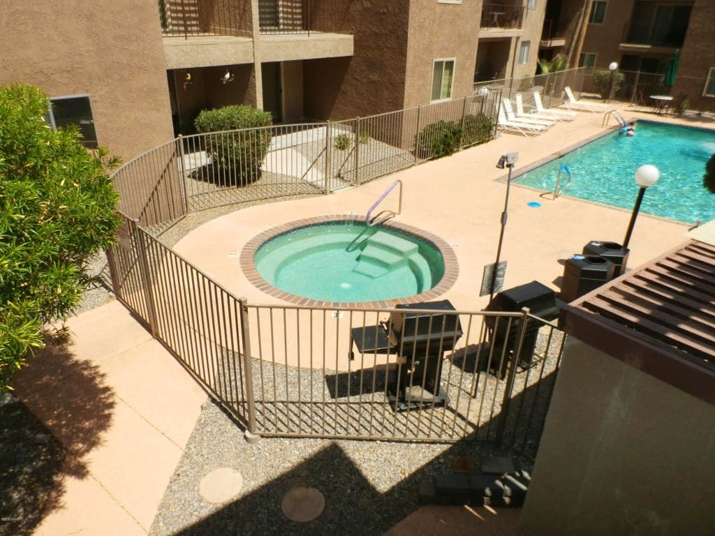 A well-maintained pool and hot tub area is enclosed by a secure fence. Sun loungers are arranged nearby on the patio. The pool features clear blue water and is surrounded by landscaped greenery, providing a relaxing outdoor space for guests.