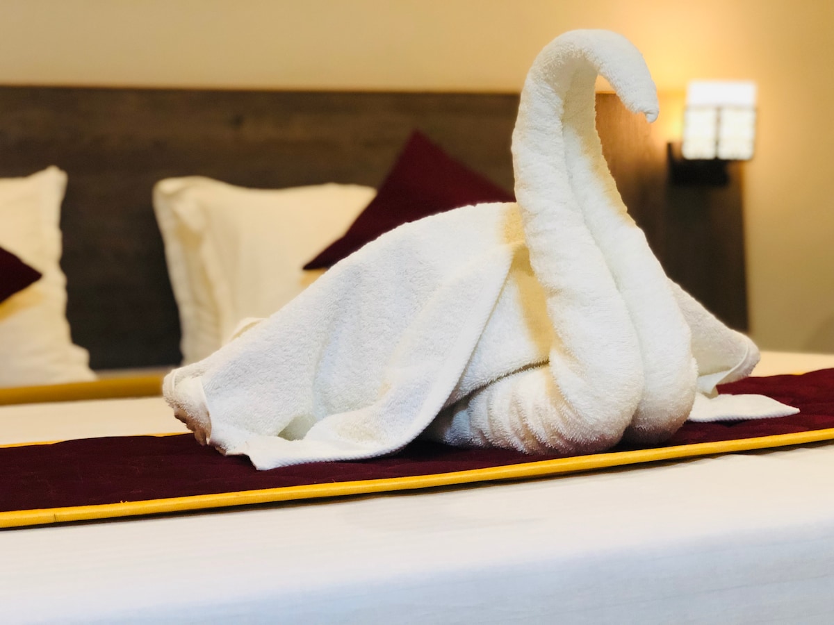 A towel sculpture in the shape of a swan is displayed on a neatly made bed with crisp linens. The headboard is dark wood, complemented by white pillows and burgundy accent cushions. Soft lighting enhances the calming atmosphere in the room.