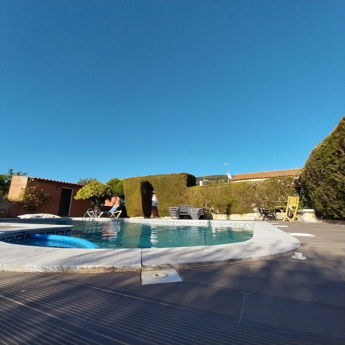 A private swimming pool area is surrounded by well-maintained hedges and greenery. Loungers are positioned nearby, inviting relaxation under the clear blue sky. A small section of the pool features a jacuzzi, offering a serene spot to unwind.