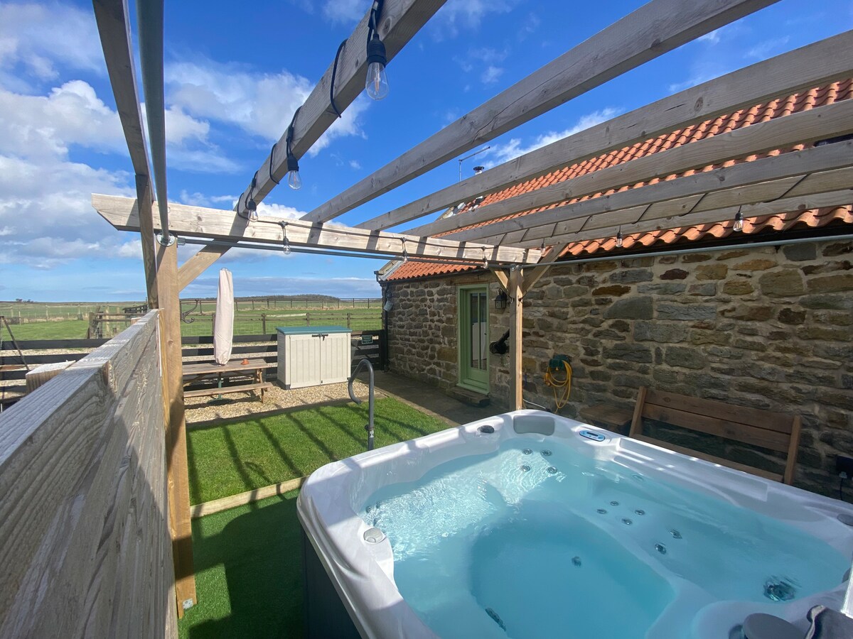 The image displays an outdoor hot tub under a wooden pergola with overhead lighting. The stone cottage is visible in the background, surrounded by green grass. A seating area and a sun umbrella can be seen nearby, with expansive views of the countryside extending beyond.