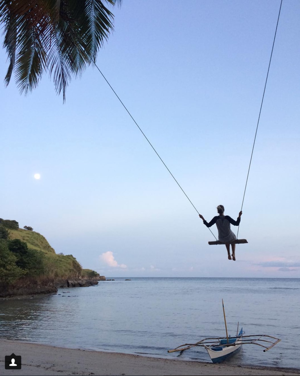 A swing hangs from two ropes, positioned along the beach near the water's edge. The calm ocean reflects soft hues of blue, with a fishing boat resting nearby. The moon is visible in the sky above, casting a serene atmosphere on the landscape.