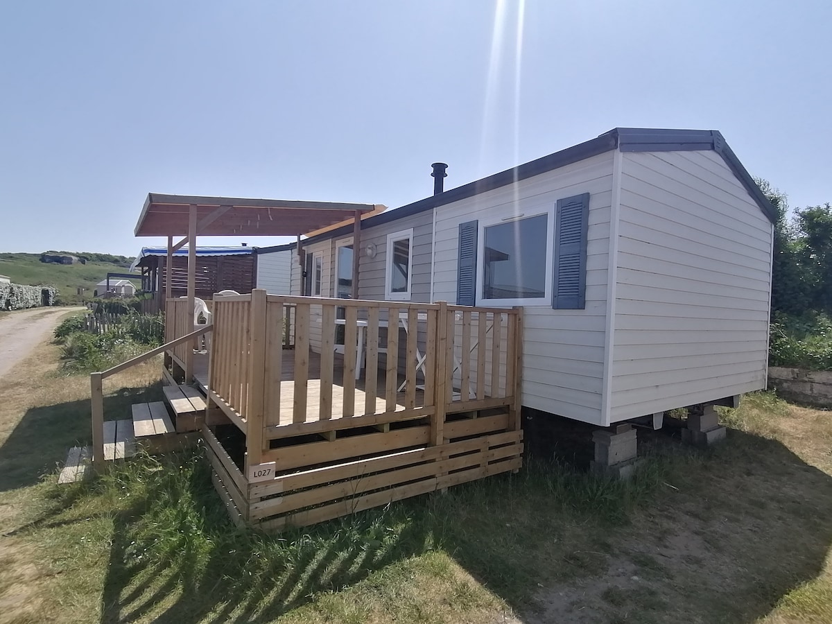 A mobile home is shown with a spacious wooden deck and an awning, situated on raised stilts. The exterior features light-colored siding and blue shutters, surrounded by grass. Nearby pathways suggest easy access to the surrounding area.