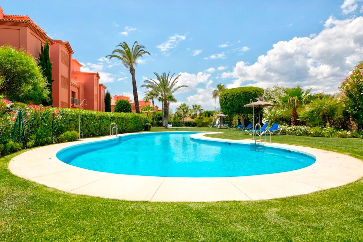 A shared swimming pool is surrounded by well-maintained gardens, featuring lush green grass and palm trees. Sun loungers are arranged near the water's edge, and a few umbrellas provide shaded areas. The sky is clear, showcasing a warm and inviting environment.