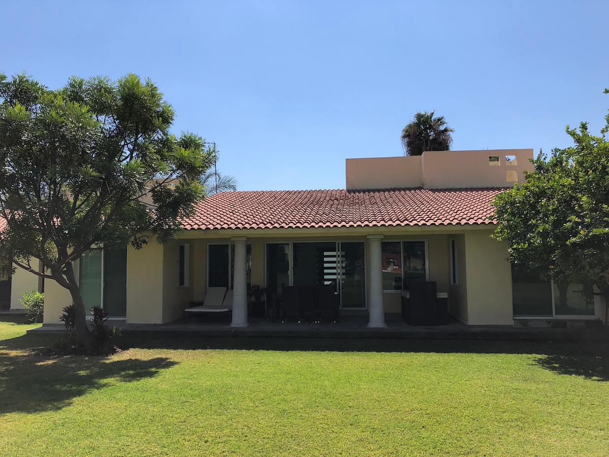 A single-story house with a tiled roof is framed by lush greenery. Large glass doors are visible, allowing ample natural light into the interior. A shaded terrace area is outlined by columns, providing a relaxing space to enjoy the surrounding garden.