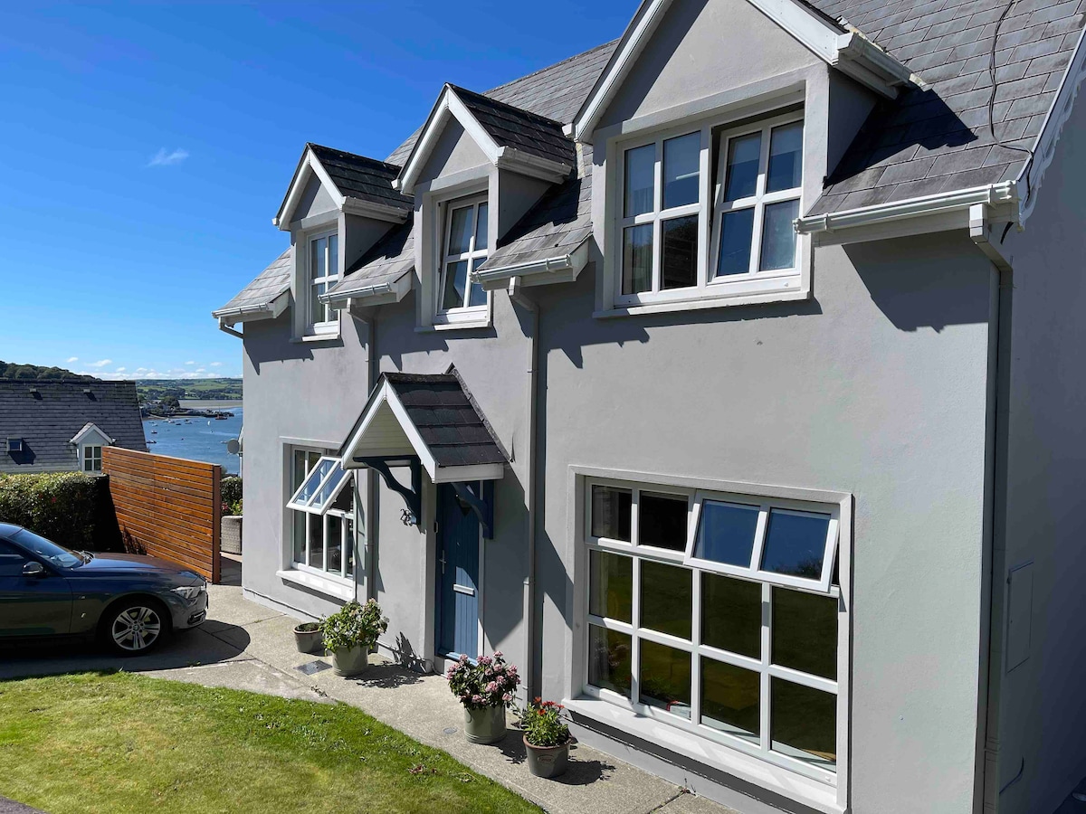 A two-story house exhibits a gray exterior with a symmetrical design. Large windows allow natural light to fill the spaces, while flower pots add a touch of greenery near the entrance. A car is parked on the driveway with a view of the bay in the background.