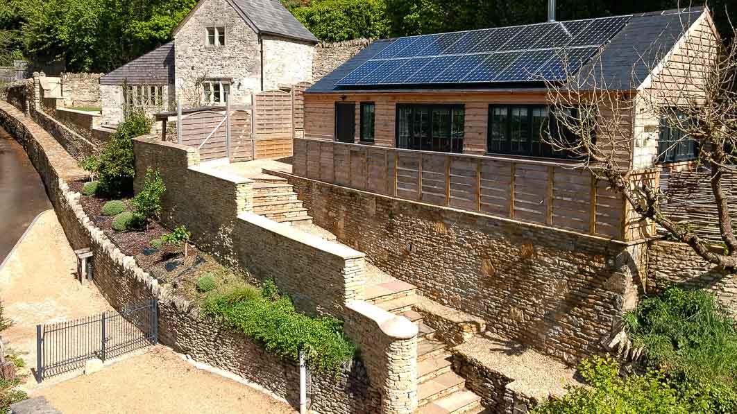 The image showcases a charming cottage nestled among greenery, constructed from stone with a modern design. Solar panels are visible on the sloped roof. A landscaped garden with neat pathways leads up to a terrace area, enhancing the outdoor space and providing a serene setting.