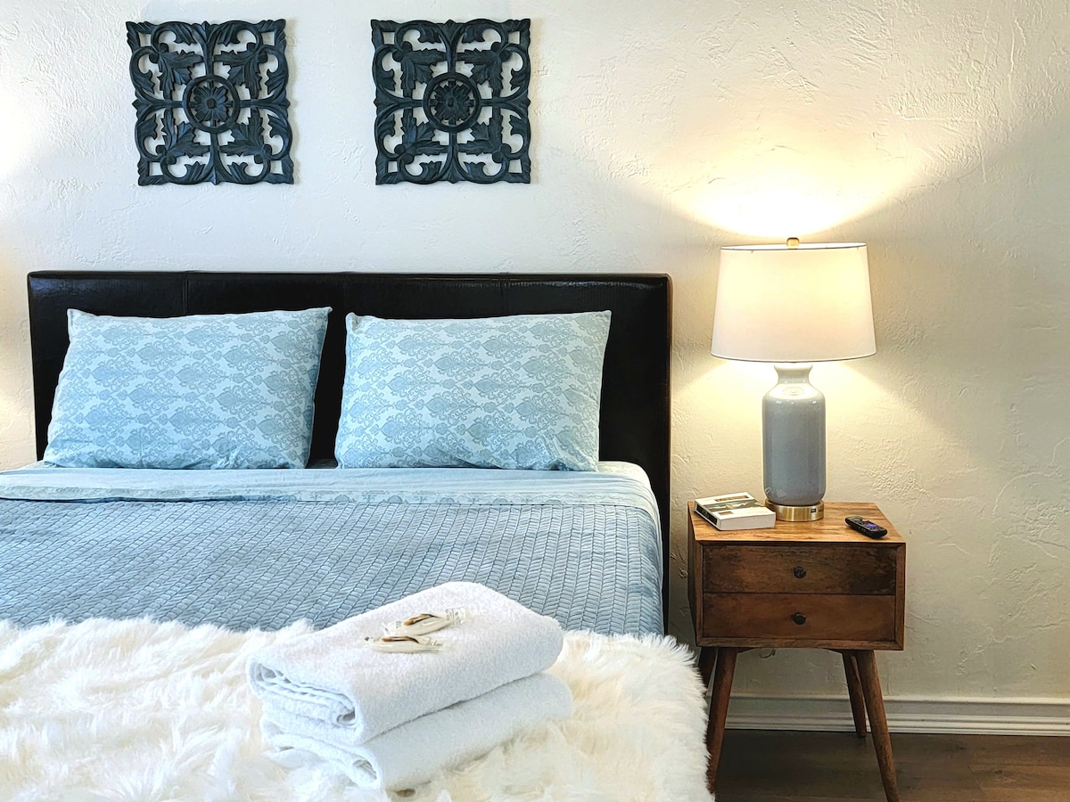A restful sleeping area is presented, featuring a queen-sized bed adorned with soft blue bedding. Two decorative wall hangings enhance the ambiance. A bedside table holds a lamp, providing gentle lighting, and neatly folded towels are placed in front of the bed.