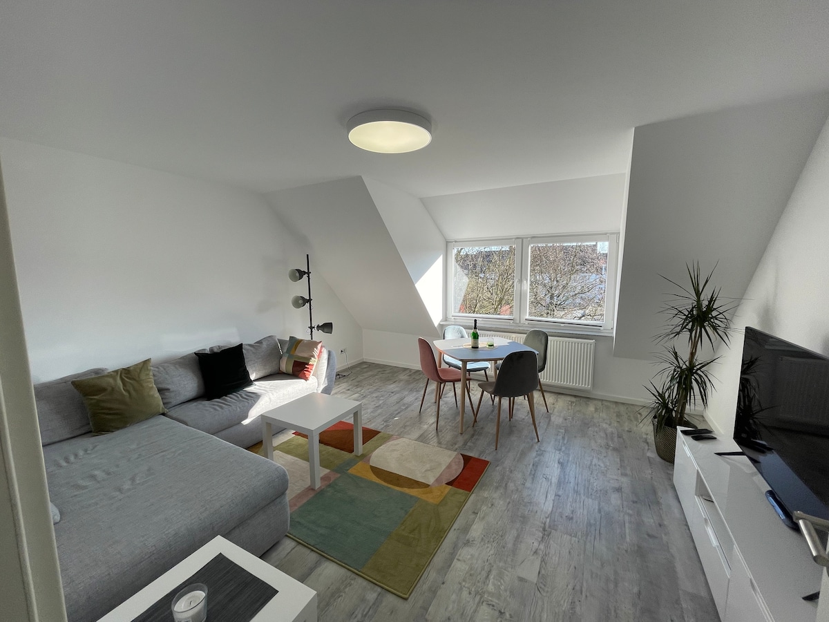 A bright living area features a sectional sofa and a minimalist coffee table. A round dining table with four chairs is positioned near large windows, allowing natural light to illuminate the space. A plant adds a touch of greenery to the corner, complementing the modern decor.