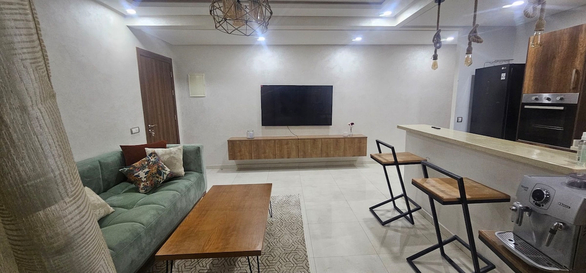 A modern living area is highlighted by a comfortable green sofa, a wooden coffee table, and stylish bar stools positioned by a dining counter. A large flat-screen television is mounted on a wooden panel, while bright ceiling lights enhance the clean, open space.