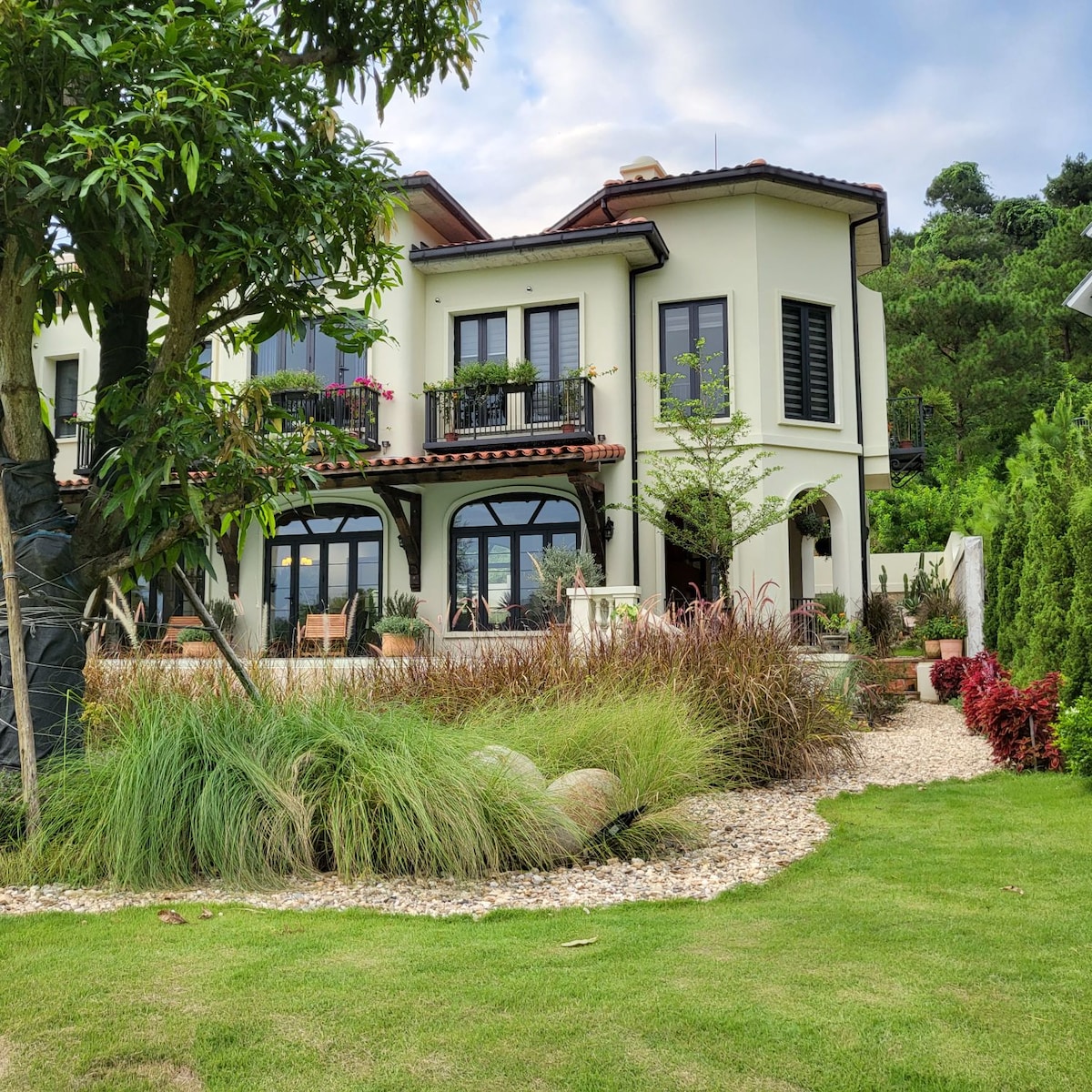The exterior of a two-story house is surrounded by manicured gardens. Lush greenery and colorful plants frame the stone pathway leading to the entrance. Large windows and balconies are present, featuring flowering plants, while a peaceful atmosphere is created by the surrounding landscape.