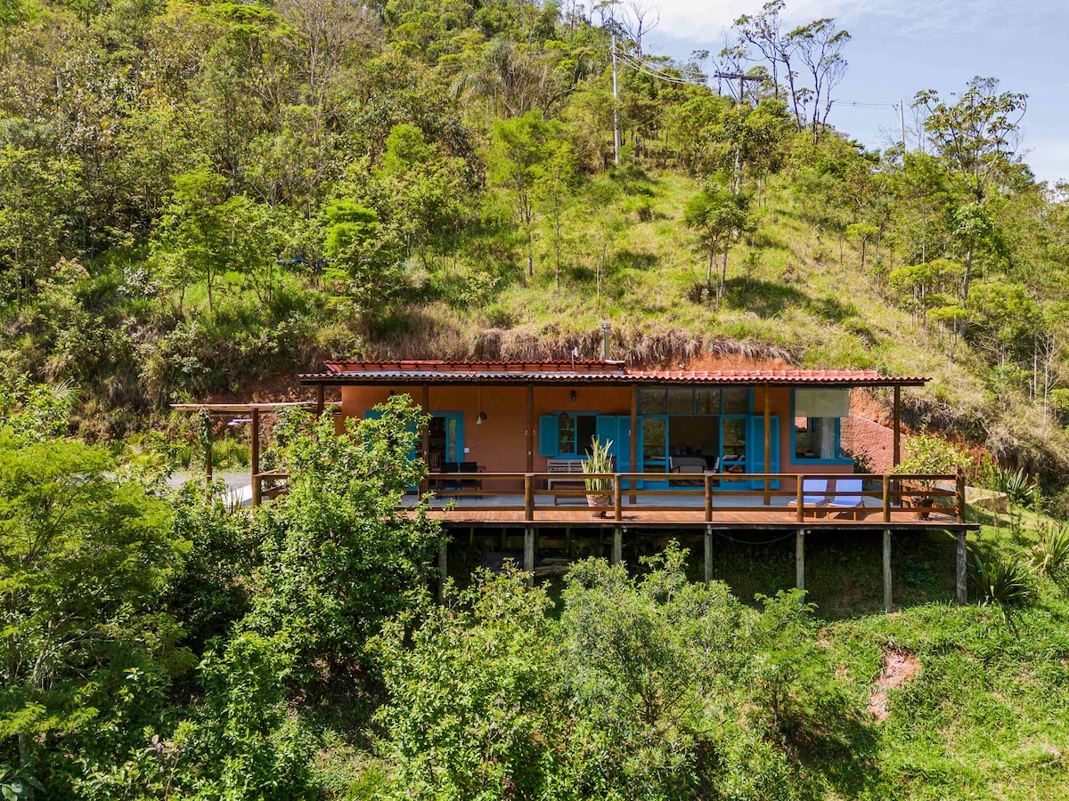 A charming house is positioned on a hillside, surrounded by lush greenery. A spacious deck extends from the front, providing opportunities for outdoor relaxation. The vibrant exterior is complemented by blue windows, contrasting with the natural landscape.