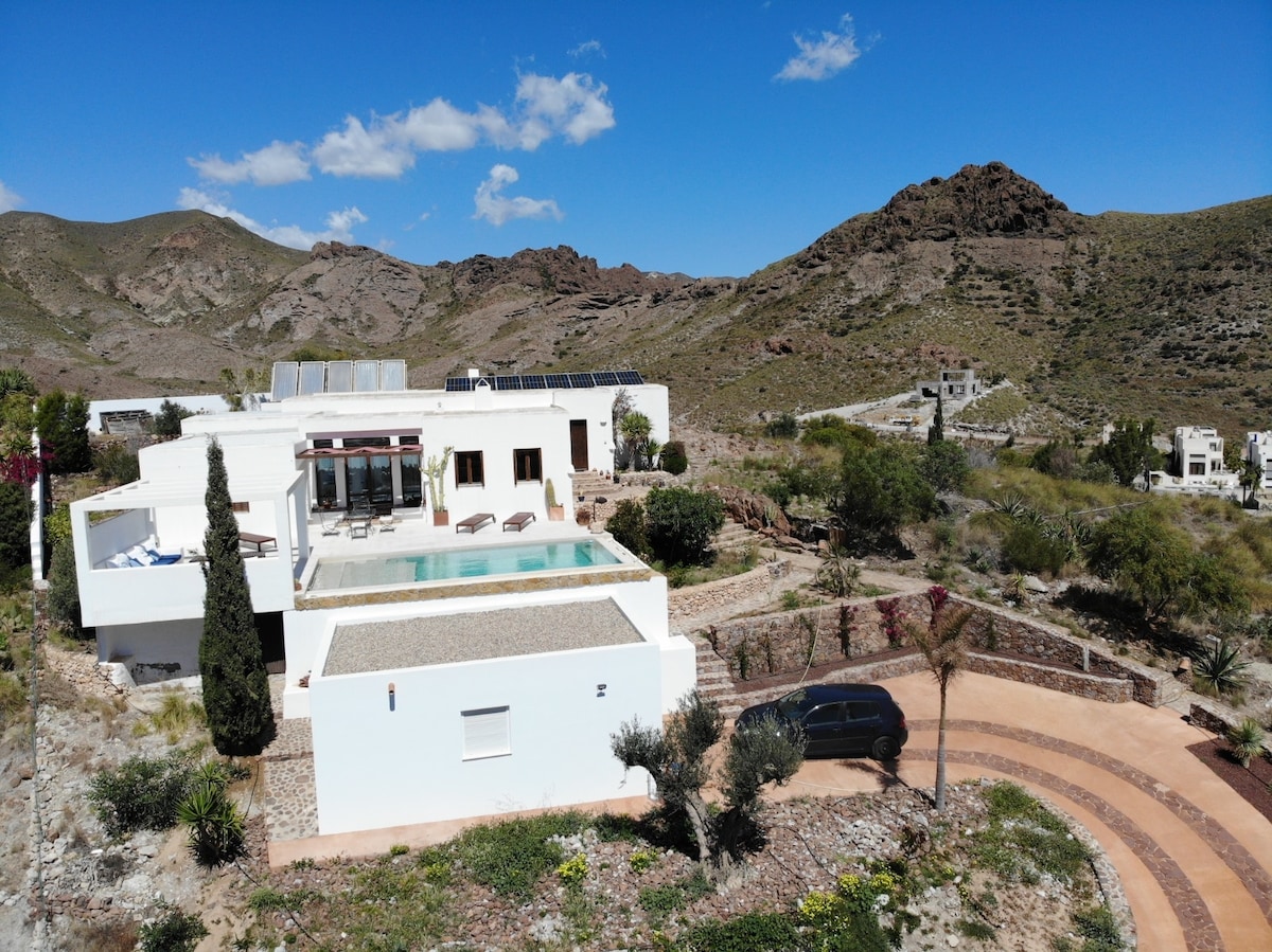 A modern house is positioned on an elevated landscape, surrounded by natural hills. An outdoor pool is visible alongside sun loungers under a clear blue sky. The driveway leads to the house, bordered by carefully arranged landscaping.