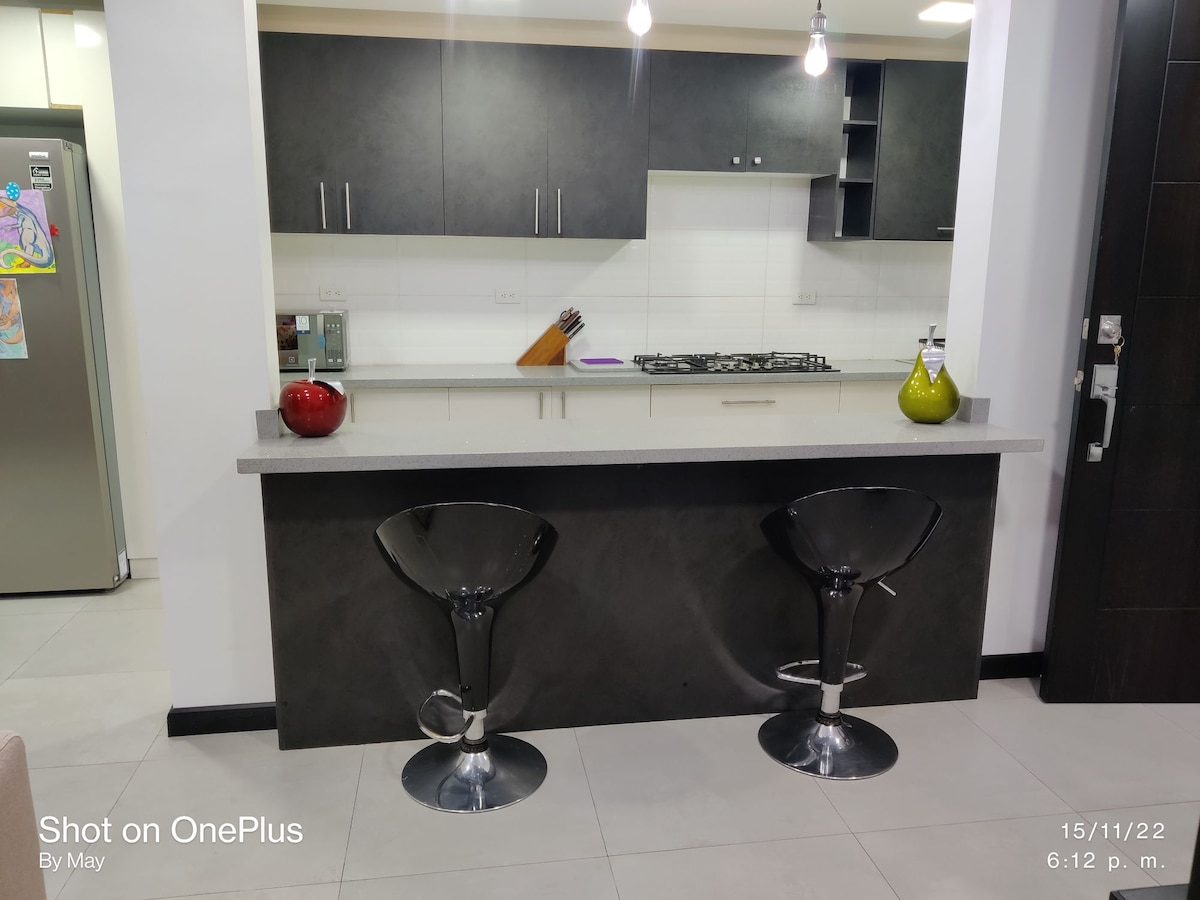 A modern kitchen area features a sleek countertop with two stylish bar stools. The kitchen is equipped with a gas stove, and storage cabinets are visible above the counter. Stainless steel appliances can be seen in the background, complemented by colorful decorative items.