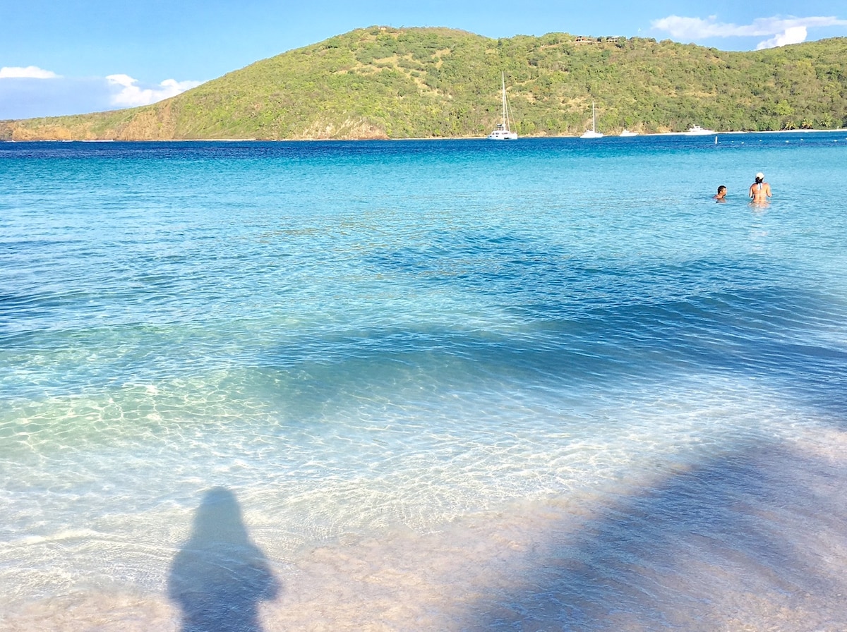 A serene beach scene features calm, clear waters reflecting shades of blue and turquoise. Gentle waves lap against a smooth sandy shore, while people enjoy the water in the distance. Lush green hills rise in the background, creating a tranquil coastal atmosphere.