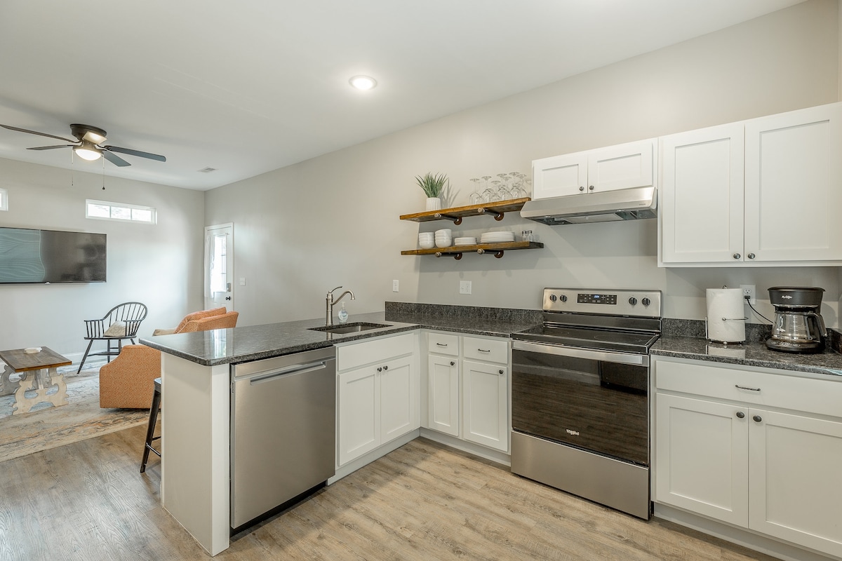 The spacious kitchen features stainless steel appliances, including a dishwasher and a coffee maker. Open shelves hold dishware, and dark countertops contrast with the white cabinetry. A living area is visible in the background, providing a comfortable seating arrangement and a smart TV.