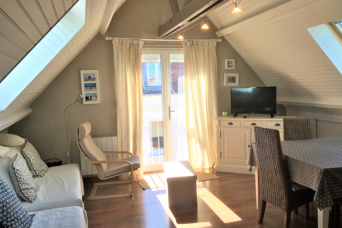 A bright living area features a comfortable sofa and a stylish armchair beside a window draped with light curtains. A television is positioned against a light-colored cabinet, while a dining table surrounded by woven chairs occupies the space. Warm light creates a welcoming ambiance.
