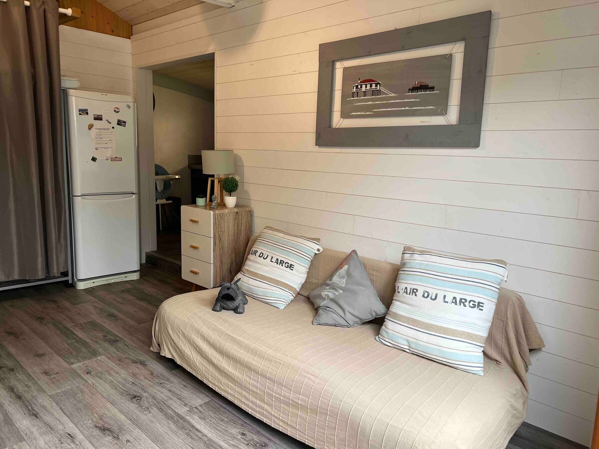 A light and airy living area features a neutral-toned sofa bed adorned with decorative pillows. A framed artwork is mounted on the white shiplap wall, and a small table is positioned next to the sofa. A refrigerator and curtain-covered doorway can be seen in the background.