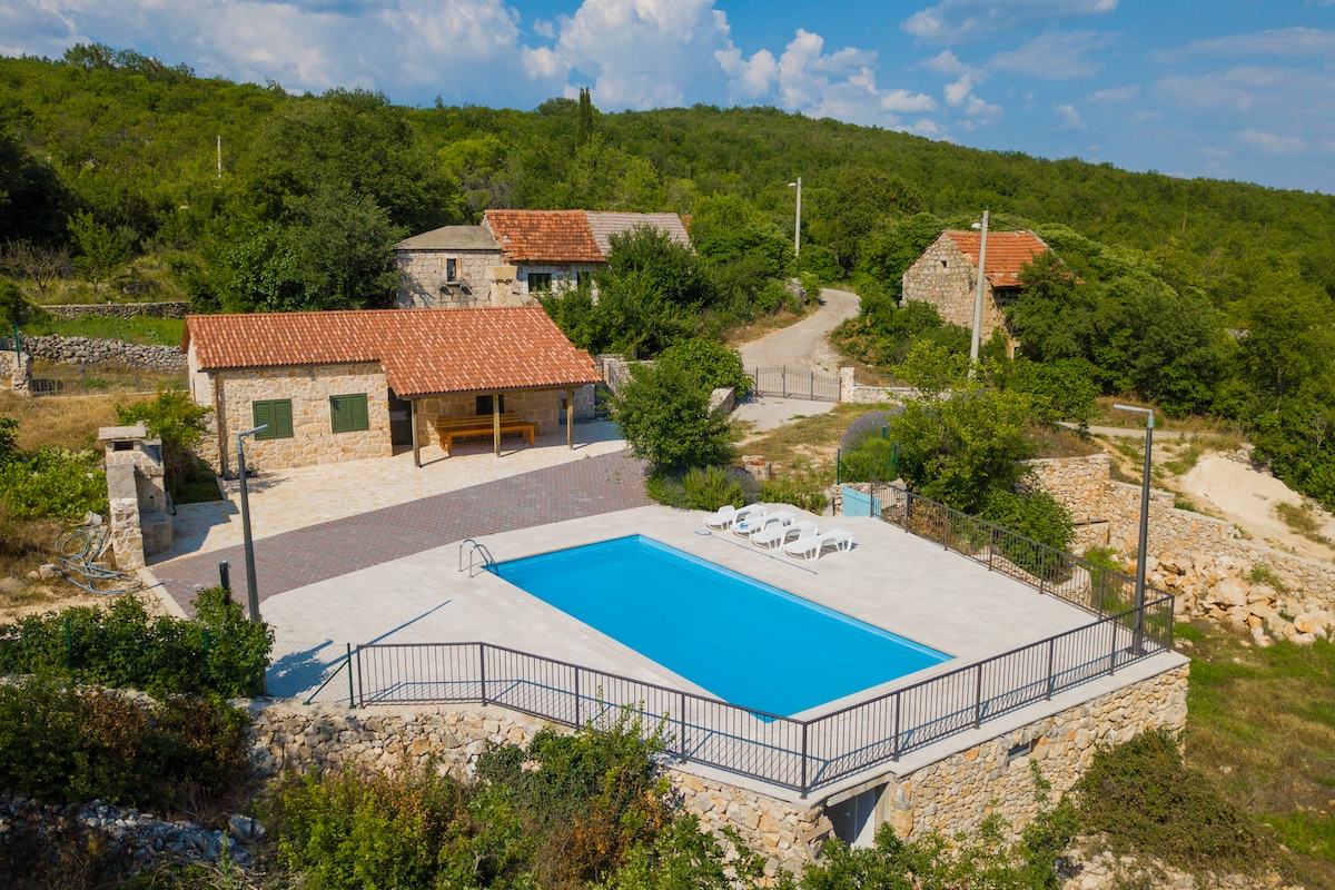 An aerial view captures a sunny outdoor area featuring a turquoise swimming pool surrounded by a spacious tiled deck. The pool area is complemented by several sun loungers and a small stone building, set against a backdrop of lush greenery and rural landscape.