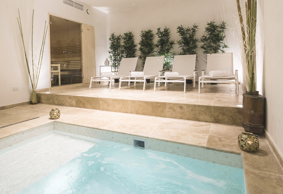 A serene spa area features a turquoise Jacuzzi, surrounded by elegant stone flooring. Four white lounge chairs are positioned nearby, accompanied by ambient lighting and lush greenery, creating a peaceful retreat.