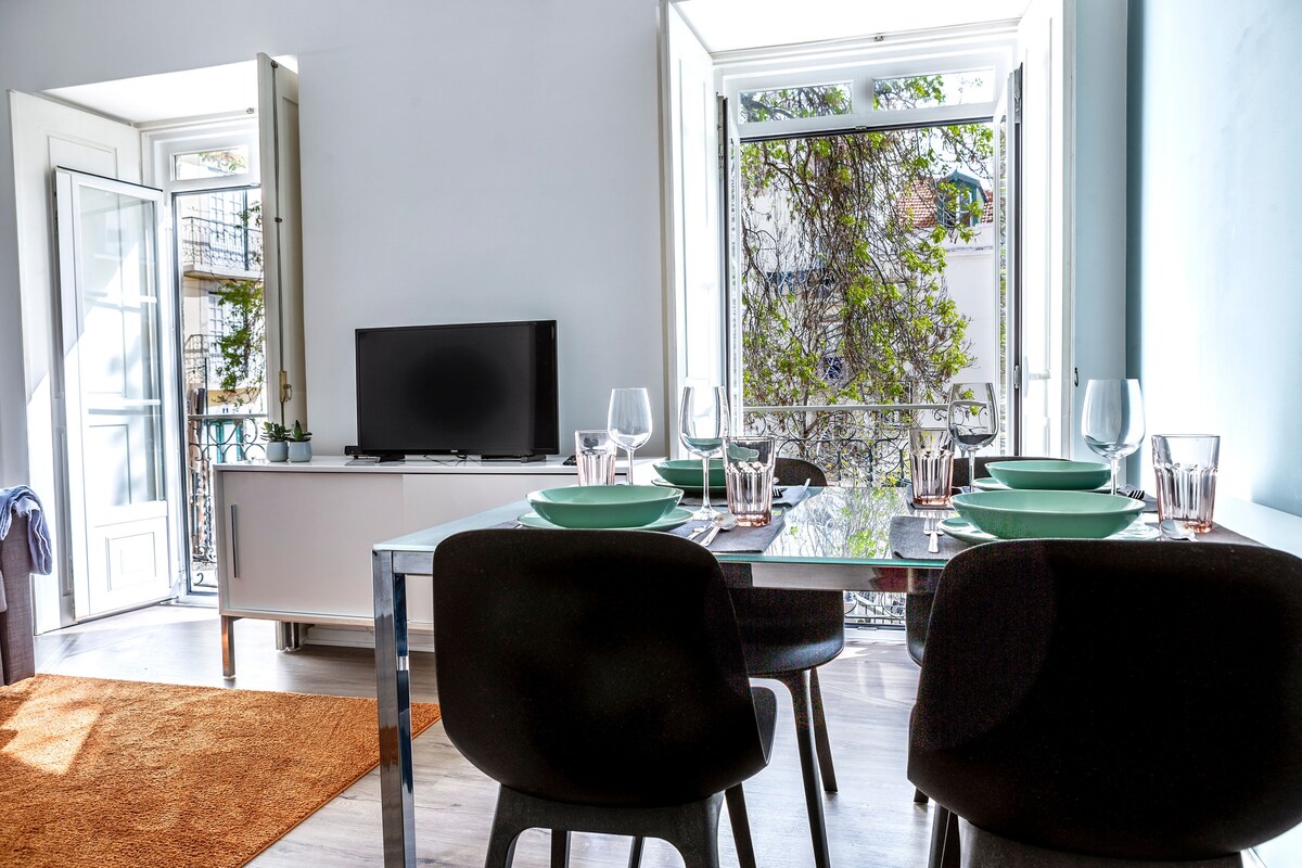A dining area is set for four, featuring a glass table with teal plates and clear glasses. Natural light floods the space through open double doors, highlighting a flat-screen TV and a cozy rug on the floor. Greenery is visible outside, creating a refreshing view.