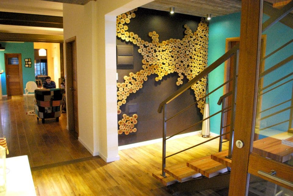 A stylish corridor showcases a unique decorative wall made of circular wooden pieces. The warm wooden floors extend throughout, leading towards an inviting seating area in the background. Modern lighting fixtures highlight the contemporary design elements, contributing to a sense of spaciousness.