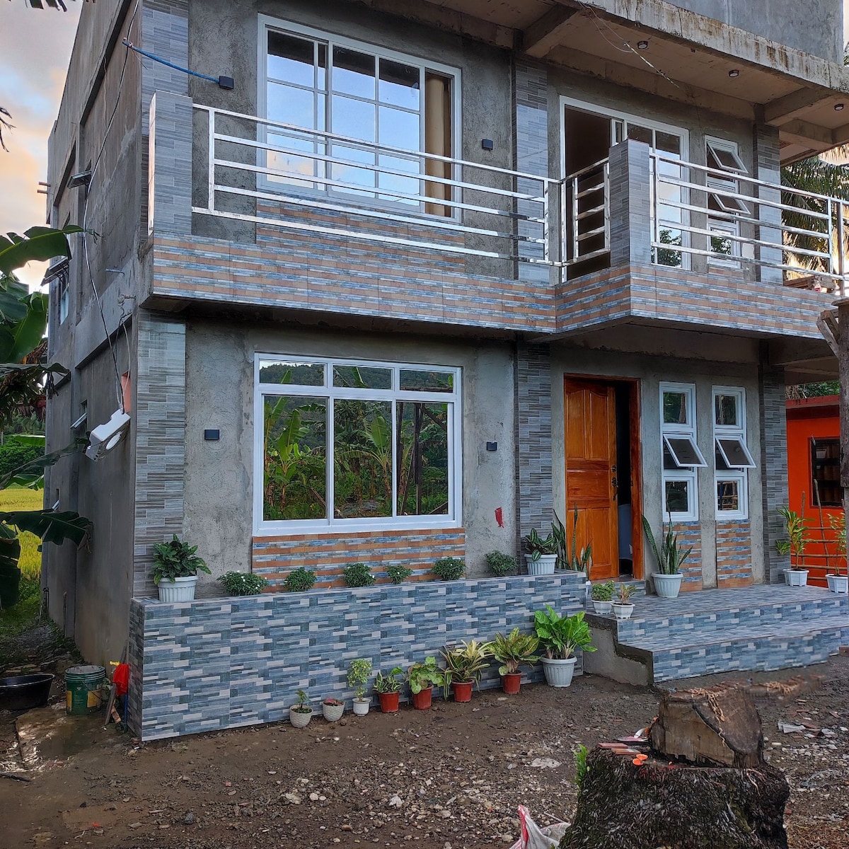 The exterior of a two-story building is shown, featuring a mix of textured stonework and bright window frames. Potted plants line the stairs leading to the entrance. Large windows allow light to enter the interior, and a wooden door provides access.