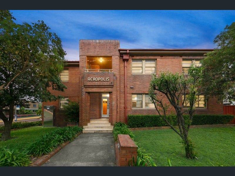 An Art Deco building is presented with rich red bricks and a decorative entrance sign reading 'ACROPOLIS.' Lush green landscaping surrounds the structure, adding a fresh element to the exterior, while soft evening light emanates from the upper windows.