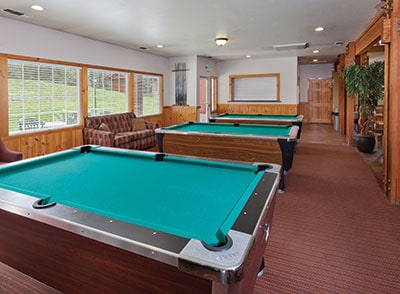 A recreation area is shown, featuring two pool tables with vibrant green felt. Natural light fills the space through large windows, illuminating a cozy seating area with a brown sofa. Wood paneling adds warmth to the interior, creating a welcoming atmosphere for leisure activities.