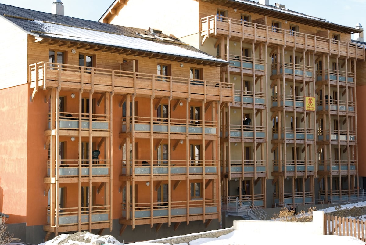 The exterior of a multi-story building is presented, featuring a blend of wooden balconies and orange-hued walls. Multiple windows are visible, contributing to a welcoming ambiance. The architecture reflects a modern alpine design, harmoniously integrating with the surrounding snowy landscape.
