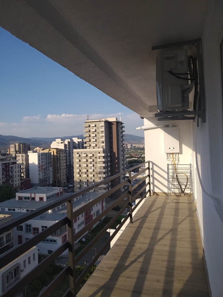 A balcony view reveals a cityscape featuring a mix of residential and commercial buildings. A railing separates the outdoor space from the inside. An air conditioning unit is mounted above, while a gentle breeze can be envisioned through the open expanse.