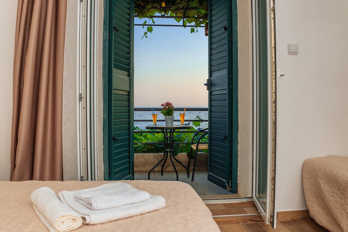 A pair of open green shutters frame a balcony overlooking the sea. A small table and two chairs are positioned on the balcony, set against the backdrop of a sunset sky. Soft linens are neatly arranged on the spacious bed.