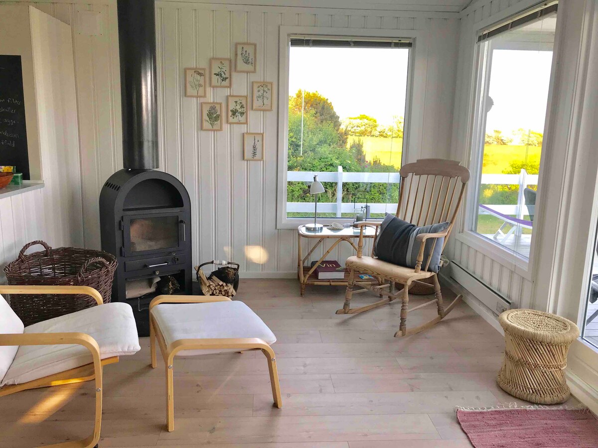 A bright and airy sitting area is created with large windows offering views of the surrounding landscape. A black wood stove stands against the white shiplap wall. A rocking chair, a small table, and a basket are arranged on the light wood floor.