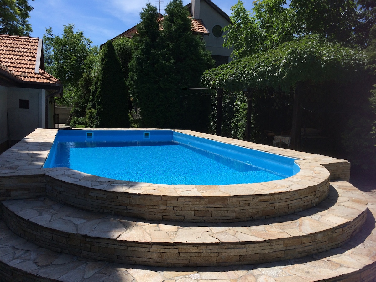 A rectangular swimming pool is surrounded by a stone terrace with tiered levels. Lush greenery frames the pool area, and a pergola provides shade nearby. The water appears clear and inviting under the sunlight, enhancing the tranquil outdoor environment.