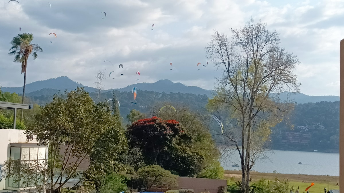 A scenic view showcases a lake surrounded by mountains, with colorful parachutes dotting the sky. Lush trees frame the landscape, providing depth to the tranquil setting. The clouds cast soft shadows over the water, enhancing the serene atmosphere.