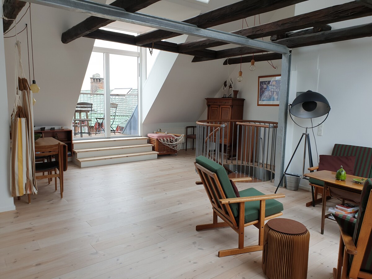The spacious loft area is filled with natural light, highlighting wooden beams and light-colored flooring. A seating arrangement includes green armchairs and a wooden table. Two steps lead to a balcony, while a cabinet and additional seating are visible in the background.