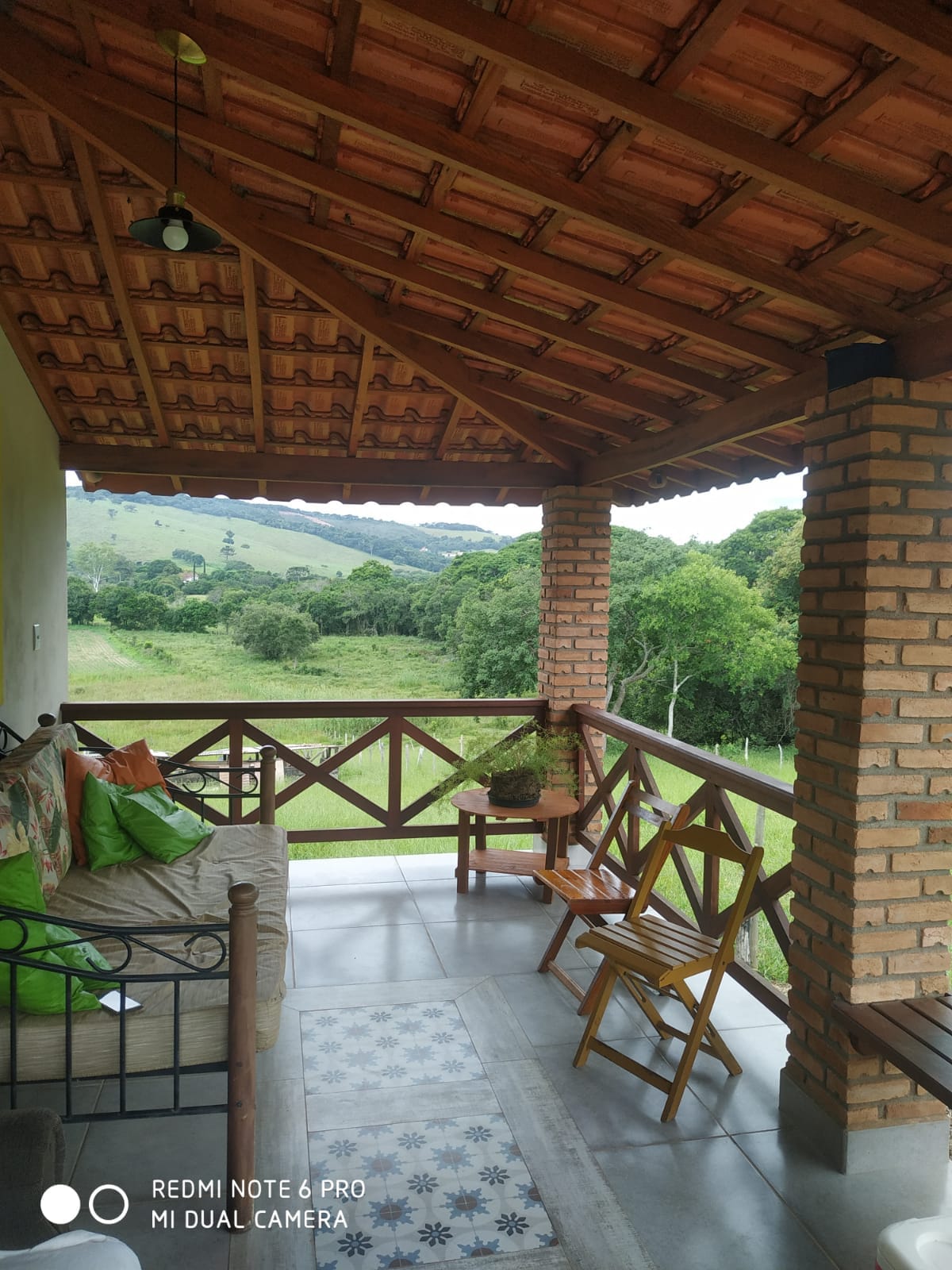 A spacious porch with a tiled floor features a comfortable seating area, including a couch and wooden chairs. The rustic brick columns support a tiled roof, while views of lush green hills and trees are visible in the background.
