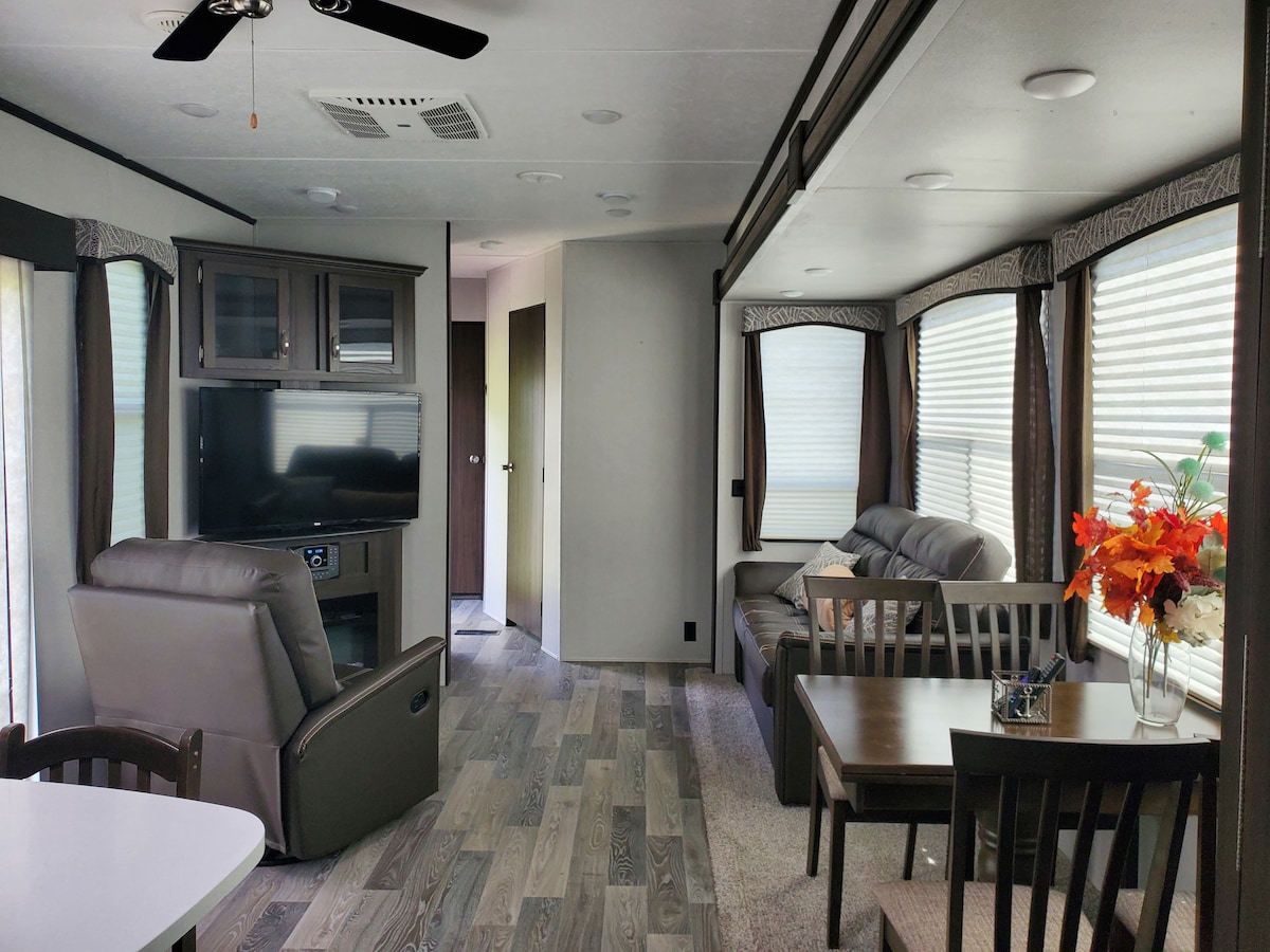 The interior of the RV features an open living area with large windows allowing natural light to fill the space. A mounted television is positioned near a comfortable recliner and a sofa. A dining table with four chairs sits beside a window, enhancing the communal space.