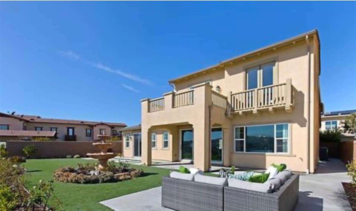 The exterior of the two-story home showcases a well-maintained garden area with a green lawn and a fountain. A covered balcony is seen on the upper floor, while comfortable seating arrangements are provided on the patio, inviting relaxation in the outdoor space.